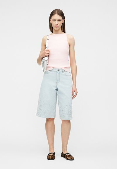 Γυναίκα που φοράει αμάνικο τοπ σε απαλό ροζ χρώμα, ανοιχτό μπλε τζιν σορτς μέχρι το γόνατο με διακοσμητικά στοιχεία, μπαλαρίνες με λεοπάρ σχέδιο και κρατάει μια ασημί τσάντα χειρός.