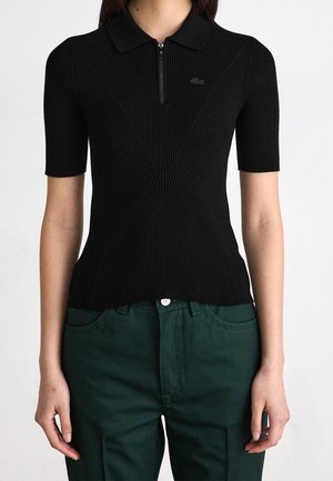 Femme portant un polo noir côtelé à manches courtes avec une fermeture éclair devant et un pantalon vert sur un fond uni.