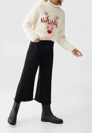 Femme portant un pull blanc pelucheux avec un renne souriant et le texte « Ho Ho Ho », associée à un pantalon noir large et court et à des bottes noires.