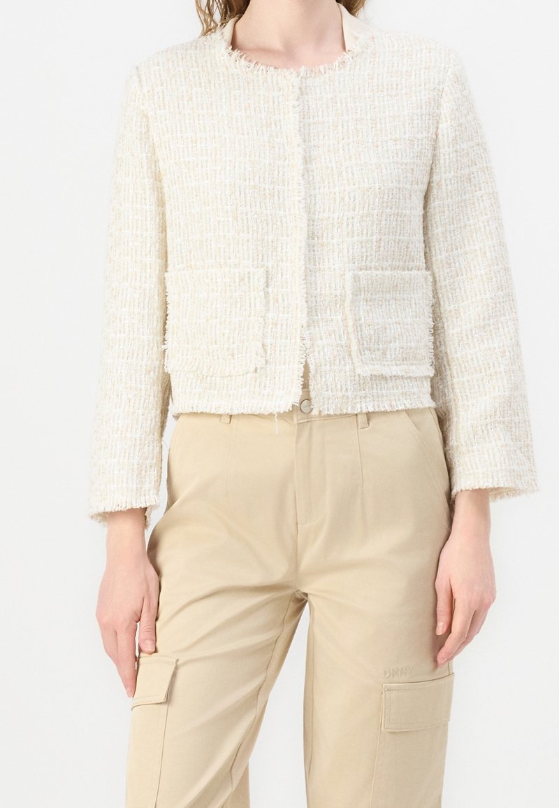Femme portant une veste courte texturée crème avec poches avant et un pantalon cargo beige avec poches à rabat, debout, bras détendus.