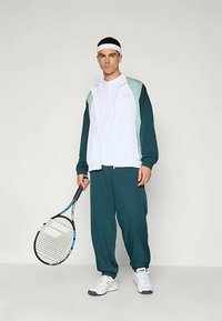 Vyrų sportinis kostiumas mėlyna ir balta. Apima užtrauktuku užsegamą striukę su plačiomis rankovėmis ir elastingomis apykaklėmis, derinamas su atitinkančiomis laisvo kirpimo kelnėmis. Laiko teniso raketę.