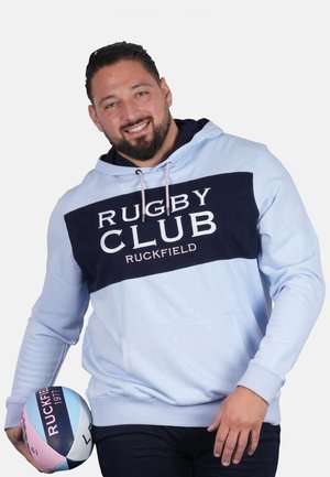 Homme souriant, portant un sweat à capuche bleu clair marqué "RUGBY CLUB RUCKFIELD", tenant un ballon de rugby aux couleurs pastel dans sa main gauche.