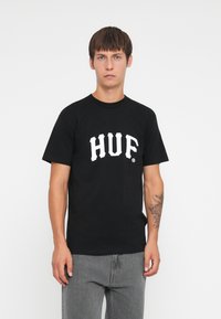 Fekete pamut póló, a mellkason feltűnő fehér "HUF" logóval. Klasszikus kerek nyakú dizájn, rövid ujjak és laza fit.