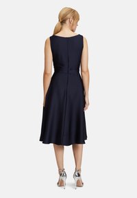 Robe A-line marine avec un col en U, sans manches et une fermeture éclair dans le dos. Une paire de sandales à talons hauts argentées.