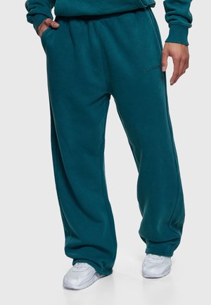 Persoon draagt turquoise joggingbroek en bijpassend sweatshirt met handen in de zakken, gecombineerd met witte sneakers, staand tegen een effen achtergrond.