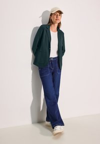 Blazer verde oscuro con dos bolsillos delanteros sobre una camiseta blanca, combinado con pantalones vaqueros azules con costuras claras y un botón verde, zapatillas blancas.