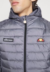 Mand iført en grå quiltet hættetrøje med sort lynlås og et hvidt "ellesse" patch på venstre bryst samt et halvcirkelformet logo på højre side.