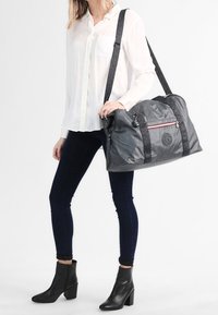 Šedá duffel taška s černými vzorovanými popruhy, zipovým uzávěrem a detailem loga. Měkký, lehký materiál s prostorným designem.