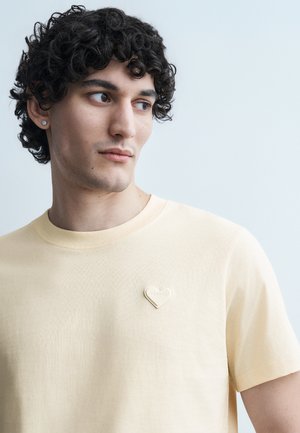 Ung mand med krøllet mørkt hår iført en beige t-shirt med en lille hjerteformet nål på venstre bryst, der ser til siden.