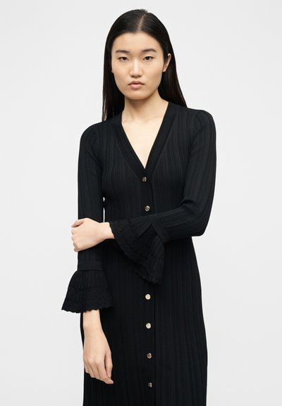 Γυναίκα με ίσια σκούρα μαλλιά που φοράει μαύρο ριμπ φόρεμα με κουμπιά στο μπροστινό μέρος και φαρδιές δαντελωτές μανσέτες, στέκεται μπροστά σε απλό φόντο.