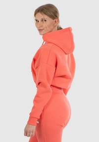 Felpa con cappuccio color corallo e pantaloni abbinati in un morbido tessuto. La felpa ha un grande cappuccio e spalle basse; i pantaloni hanno una vita elasticizzata.