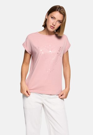 Mujer con una camiseta rosa con diseño de mariposa y estrella, combinada con pantalones blancos, posando contra un fondo liso.
