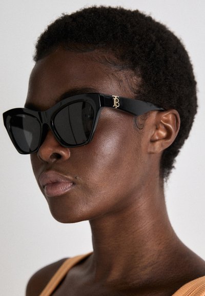 Gafas de sol negras con una forma geométrica de ojo de gato, que cuentan con lentes oscuros y acentos de logo en tono dorado prominente en las patillas negras.