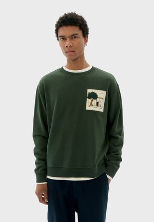 Green sweatshirt with a textured fabric, featuring a front patch with a tree design and picture frame detail. Moderate fit and crew neckline.