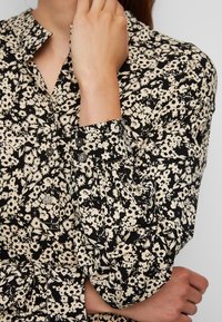 Femme portant une chemise noire à manches longues avec un motif floral crème, main touchant le visage, l'autre bras croisé à la taille.