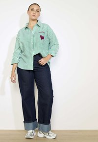 Groen-wit gestreepte shirt met een zak en een ontwerp van een rode hart, gecombineerd met donkerblauwe wijdvallende jeans en zilveren sneakers.