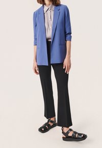 Modrý blazer s vykasanými rukávy, světle šedá košile s límcem, černé rozšířené kalhoty a černé sandály na pásku. Hladké látky, jednoduchý design.
