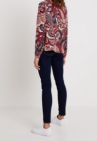Langarm-Paisleymuster-Bluse mit einem cremefarbenen Hintergrund und roten, blauen und marineblauen Mustern; kombiniert mit dunklen Skinny-Jeans und weißen Sneakern.