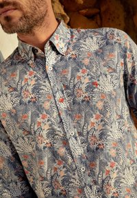 Overhemd met knopen, voorzien van een blauw-wit bloemenpatroon met oranje accenten. Gemaakt van stof met een gladde textuur en een klassieke kraag.