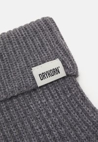 Sivý pletený čiapka s rebrovanou textúrou, s bielou etikettou s nápisom "DRYKORN" v čiernom písmene na manžete.