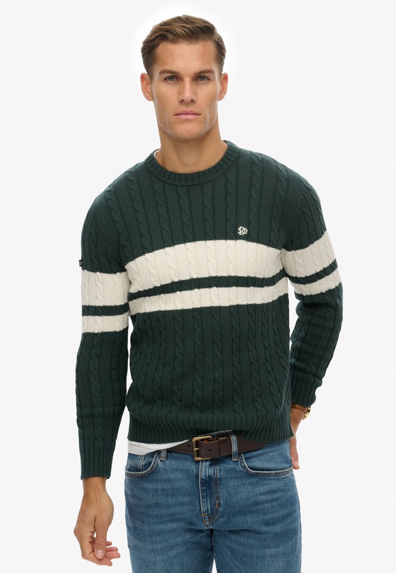 Maglione a trecce verde scuro con una striscia orizzontale bianca, scollatura rotonda e logo sul petto. Indossato con jeans blu e una cintura marrone.