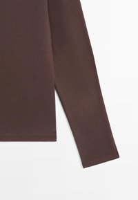 Mouw van een donkerbruin, effen katoenen overhemd met lange mouwen en zijzoom, afgebeeld op een witte achtergrond.