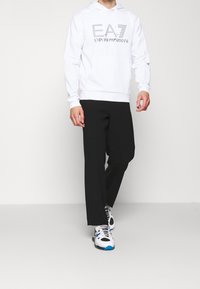 Bílá mikina s kapucí, která má šedé logo "EA7 Emporio Armani", spárovaná s černými zúženými kalhotami a bílými teniskami s modrými akcenty.