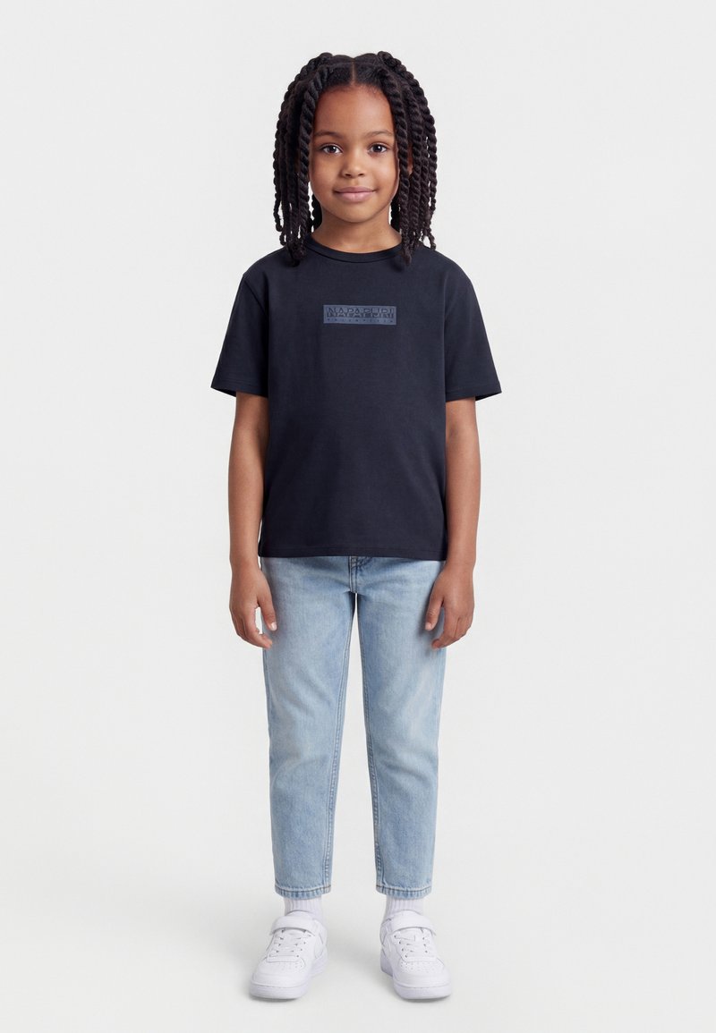 Νεαρή κοπέλα με πλεξούδες, φοράει μαύρο T-shirt με λογότυπο, ανοιχτό μπλε τζιν, λευκές κάλτσες και λευκά αθλητικά παπούτσια, στέκεται στραμμένη προς τα εμπρός.