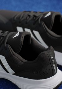 Ζευγάρι μαύρα και λευκά αθλητικά παπούτσια Adidas Gamecourt 2 σε μπλε επιφάνεια, με λωρίδες στο πλάι και λεπτομέρειες στη φτέρνα.