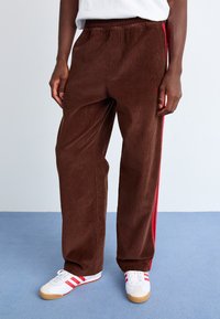 Brown corduroy pants with a textured finish, featuring red side stripes and an elastic waistband, paired with white sneakers.
