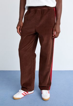 Brown corduroy pants with a textured finish, featuring red side stripes and an elastic waistband, paired with white sneakers.