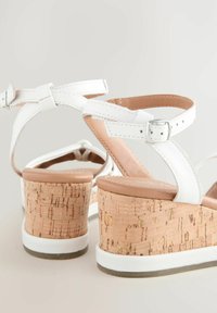 White strappy sandals with a cork wedge heel, featuring a smooth leather upper and adjustable ankle strap with a metallic buckle.