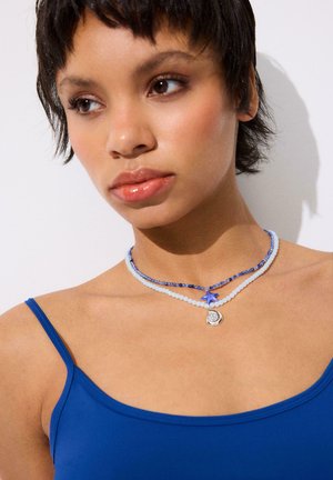 Junge Frau mit kurzem dunklem Haar trägt geschichtete blaue und weiße Perlenketten mit Seestern- und Mondanhängern, gekleidet in ein blaues Spaghettiträger-Top.