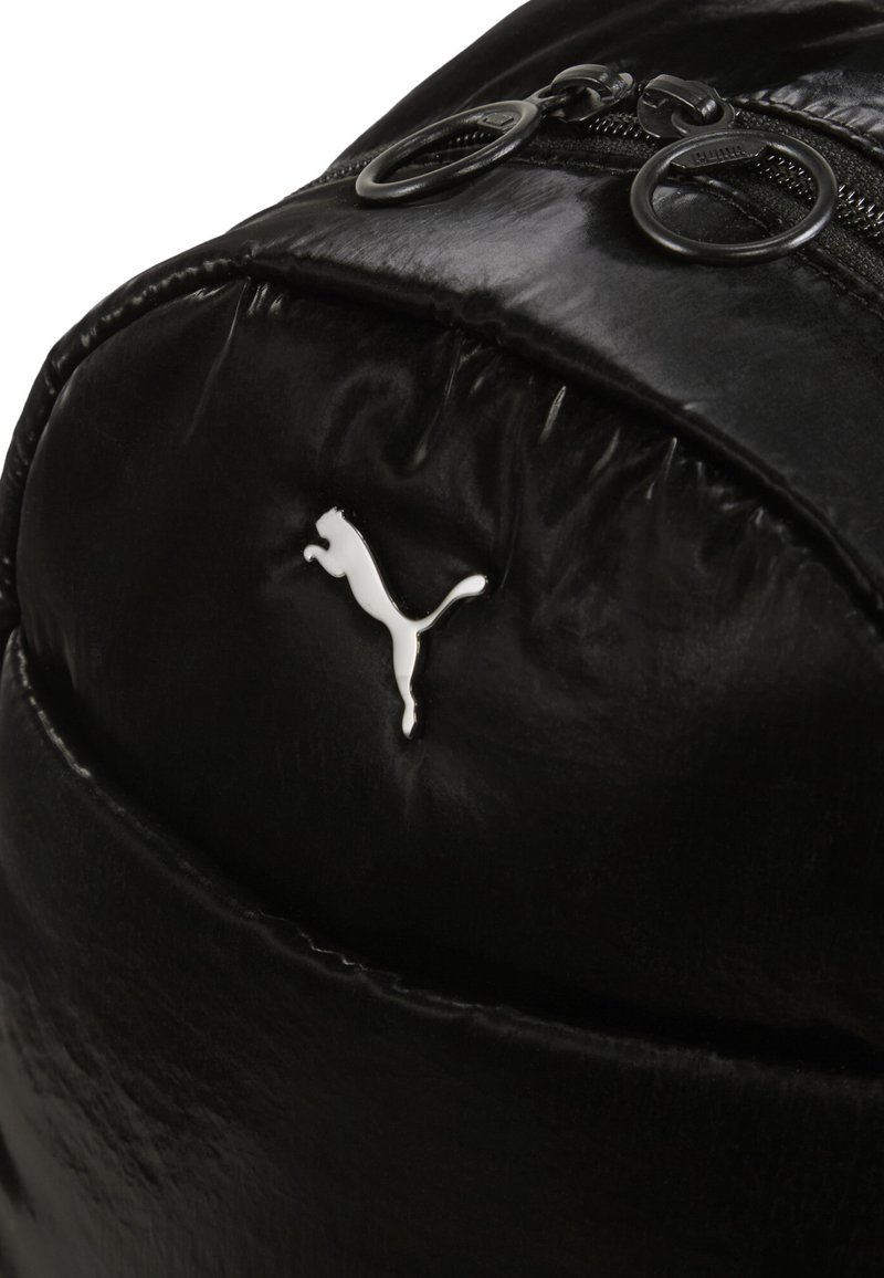 Schwarzer Leder-Rucksack mit glänzender Textur, ausgestattet mit Doppelreißverschluss-Zippern und einem weißen Puma-Logo. Gewellte Form mit glatter Oberfläche.