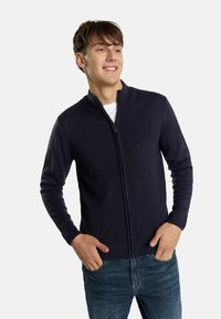 Tumši zilā krāsā zip-up džemperis ar augstu apkakli, ribotu piedurkņu un jostas daļu; izgatavots no adīta auduma, valkāts pār baltu krekla un džinsu.