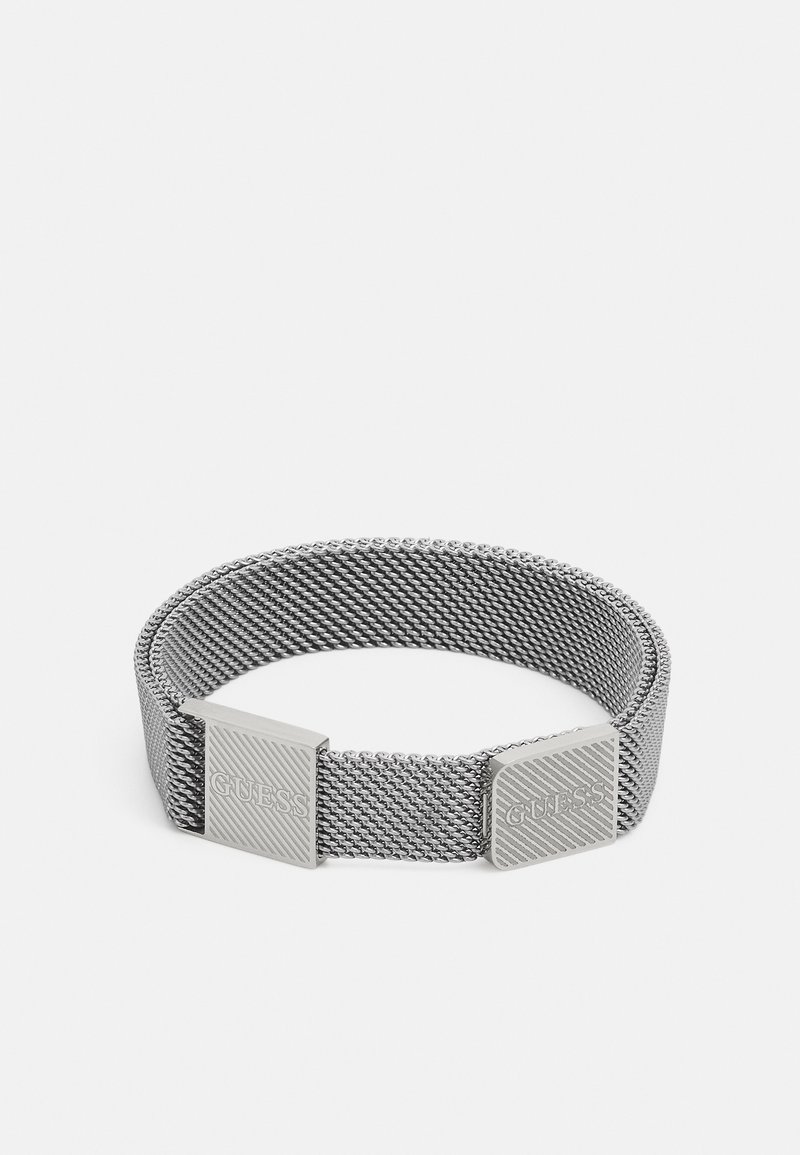 Bracelet en maille argentée avec deux fermoirs carrés gravés du nom de la marque Guess sur un fond blanc uni.