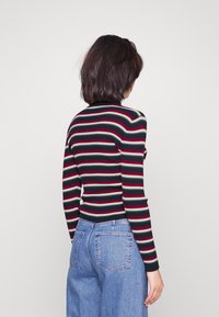 Maglione a coste con righe orizzontali di verde scuro, rosso e grigio chiaro, maniche lunghe e una vestibilità aderente; abbinato a jeans a vita alta di colore azzurro chiaro.