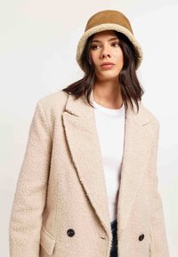 Beige boucle jas met dubbele knopen en twee voorzakken, gecombineerd met een bruine bucket hat met een pluche voering.