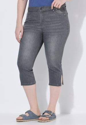 Kvinde iført grå korte jeans med sideslidser og blå slide sandaler, står op ad en hvid væg.