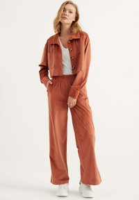 Rostfarbene Cordjacke mit Knöpfen und einer Tasche, kombiniert mit passenden weit geschnittenen Hosen, getragen mit einem weißen Oberteil und Sneakers.