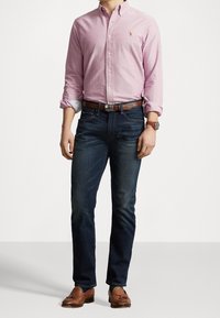 Jasnoróżowa koszula z guzikami i podwiniętymi rękawami, ciemnoniebieskie dżinsy, brązowy skórzany pasek oraz brązowe mokasyny z detalem w postaci frędzla.