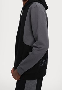 Svart och grå hoodie med en färgblockdesign, tillverkad av mjukt tyg, med snörning i huvan och sidofickor. Försedd med svag text.