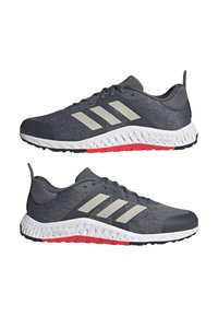Dark gray athletic shoes with a textured knit upper, three white stripes, and a dual-density white and red sole. Rounded toe and lace-up design.
