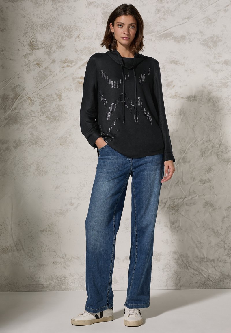 Sweatshirt à capuche noir avec des détails géométriques argentés, associé à un jean bleu à coupe décontractée et des baskets blanches avec des accents noirs.