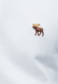 Καφέ κεντημένο αλκιονί με κίτρινα κέρατα σε λευκό υφασμάτινο φόντο με υφή, πιθανώς πουκάμισο ή υφασμάτινο αντικείμενο.