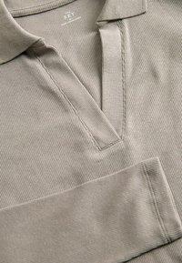 Beżowa koszulka polo z prążkowanym wzorem, o dekolcie w kształcie litery V, z teksturowanym materiałem i widocznymi detalami stębnowania na plisie i rękawach.