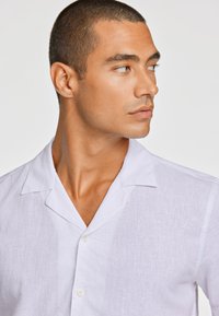 Camisa de lino blanca con diseño de cuello, de manga corta, cierre frontal de botones y un acabado texturizado, que resalta la tela transpirable.