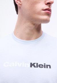 Νέος άνδρας που φοράει λευκό μπλουζάκι με το λογότυπο "Calvin Klein" σε λευκά και μαύρα γράμματα, κοιτάζοντας στο πλάι με φόντο απλό.