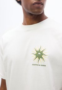 Άνδρας που φοράει λευκό μπλουζάκι με πράσινο και κίτρινο λογότυπο ηλιακής ακτίνας και το κείμενο "Scotch & Soda" στο στήθος.