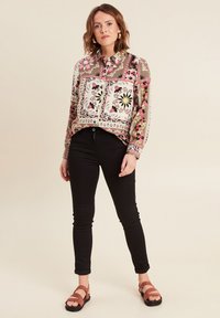 Blusa a maniche lunghe, con motivi floreali in rosa, nero e beige su uno sfondo neutro, abbinata a jeans skinny neri e sandali marroni.